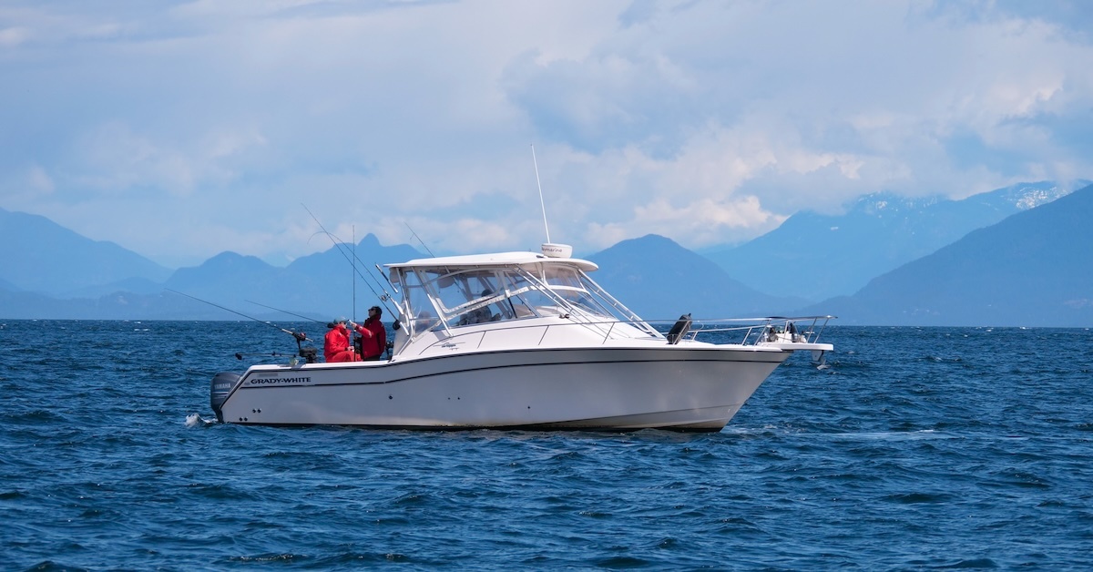 A white Grady-White walkaround fishing boat navigates the deep blue waters near Nanaimo, British Columbia. Two anglers in red waterproof gear are positioned at the stern, tending to multiple trolling lines. The backdrop features the dramatic, layered blue silhouettes of the Coast Mountains under a bright, cloud-dappled sky. Will the Salmon Allocation Policy Change this way of life?