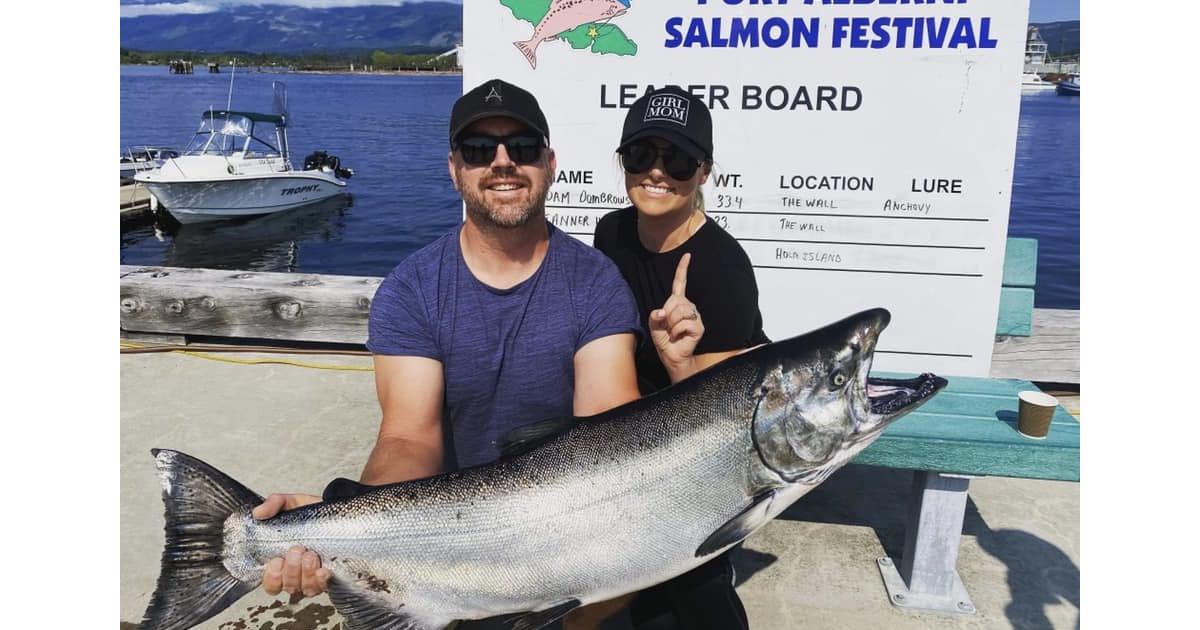 Man and woman holding a large Chinook salmon at a fishing festival, with a leaderboard in the background.
