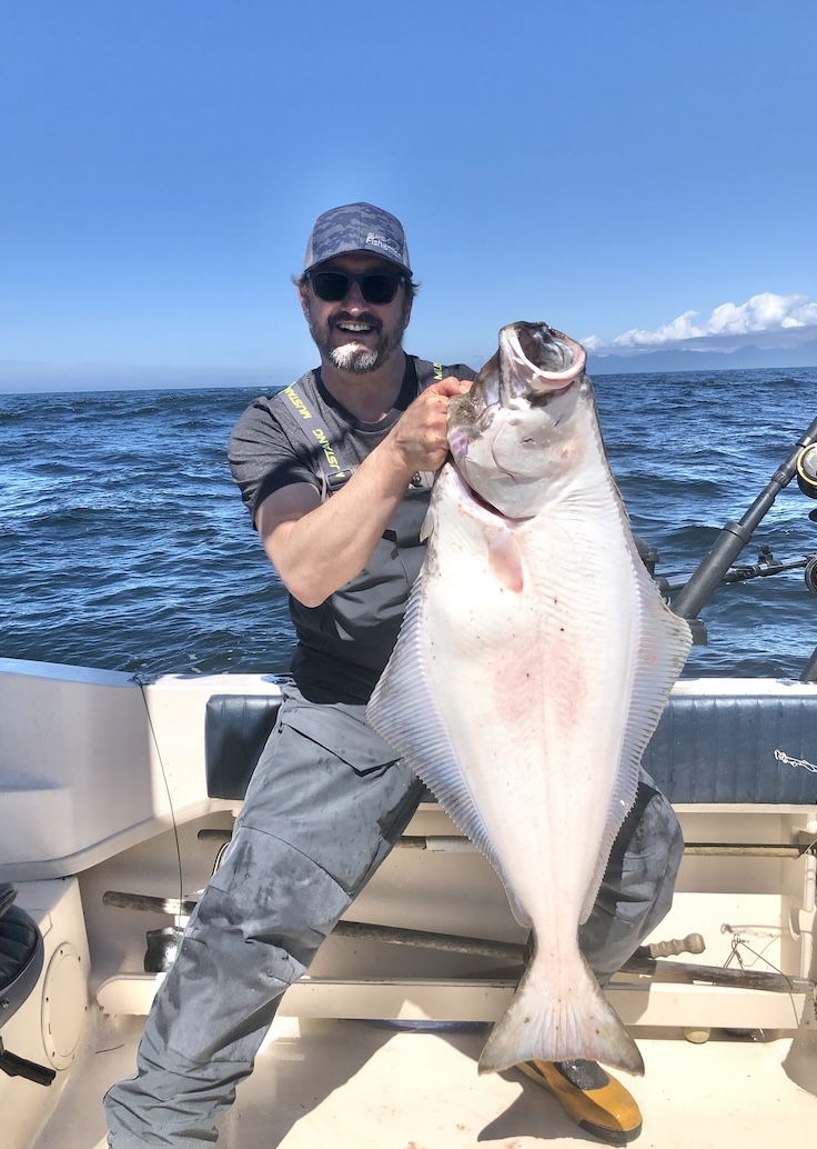 Man holding a large halibut while standing on a boat in the ocean, wearing a cap and casual fishing attire.