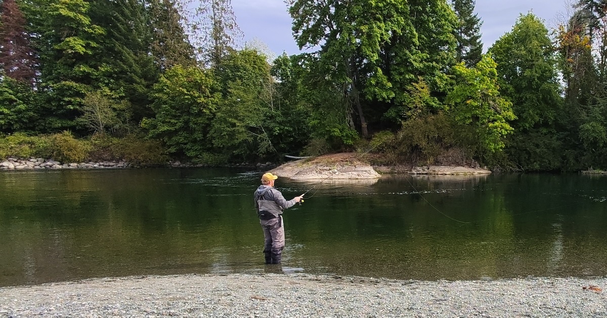 Person fishing in a river with trees and a rocky area in the background, casting a line into the water.