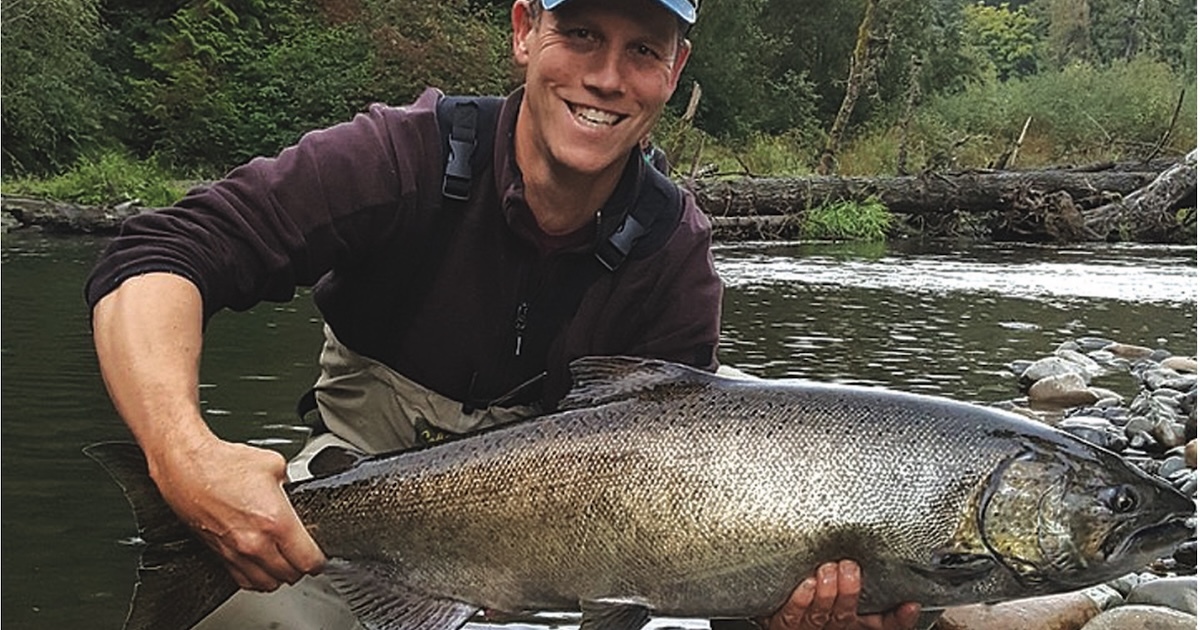 Person holding a large salmon fish while standing in a river, wearing fishing gear and a cap.