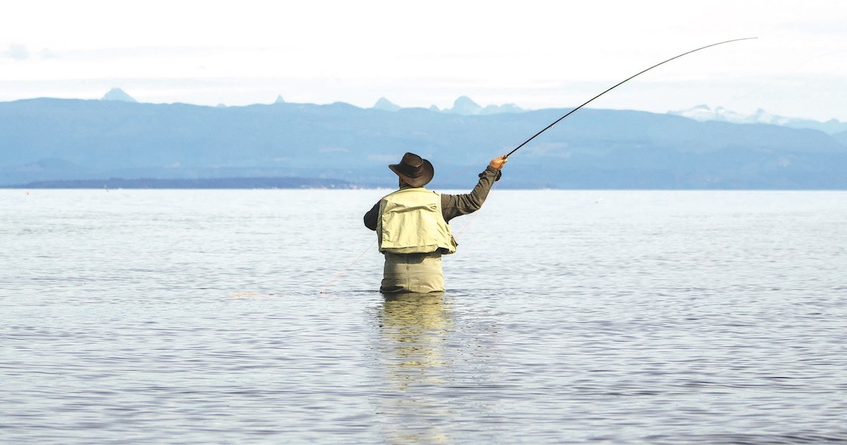 Person casting a fishing line while standing in shallow water, with mountains visible in the background.