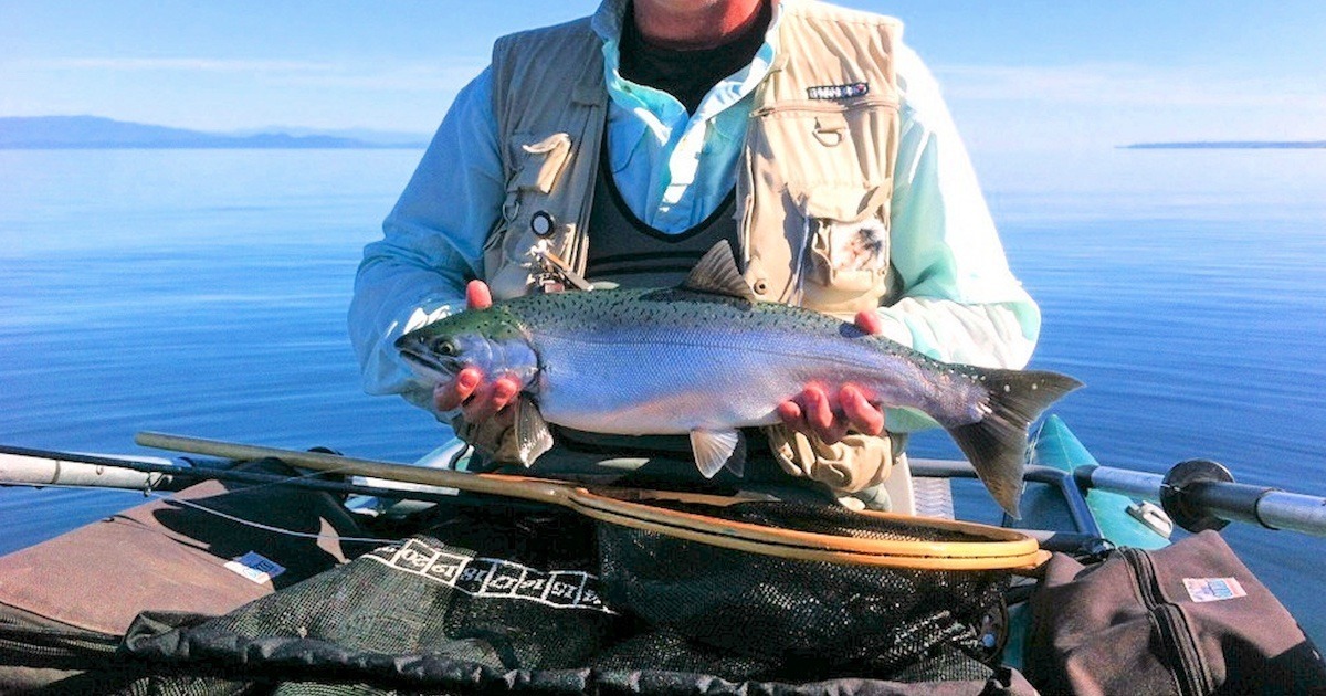 Individual holding a Coho salmon caught on a fly rod while seated in a boat on calm water.