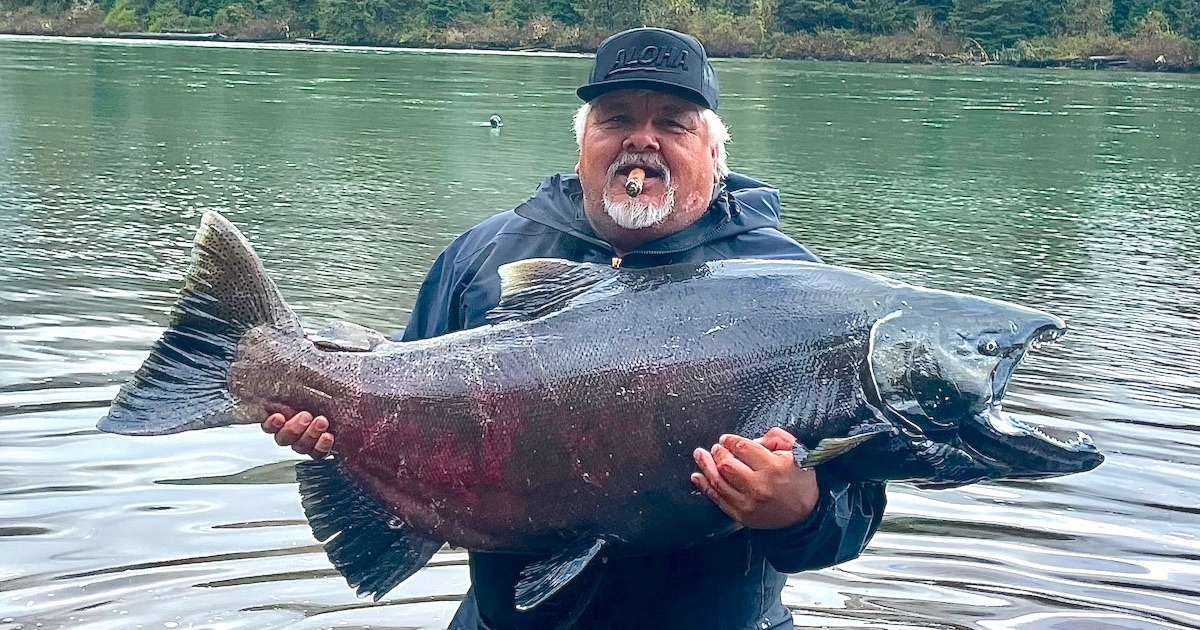 Man holding a large salmon fish while standing in water, wearing a black cap and smoking a cigar.