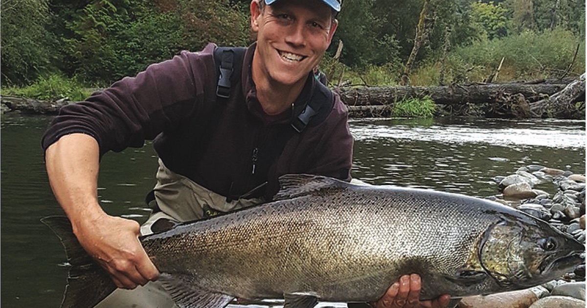Person holding a large salmon fish while standing in a river, wearing fishing gear and a cap.