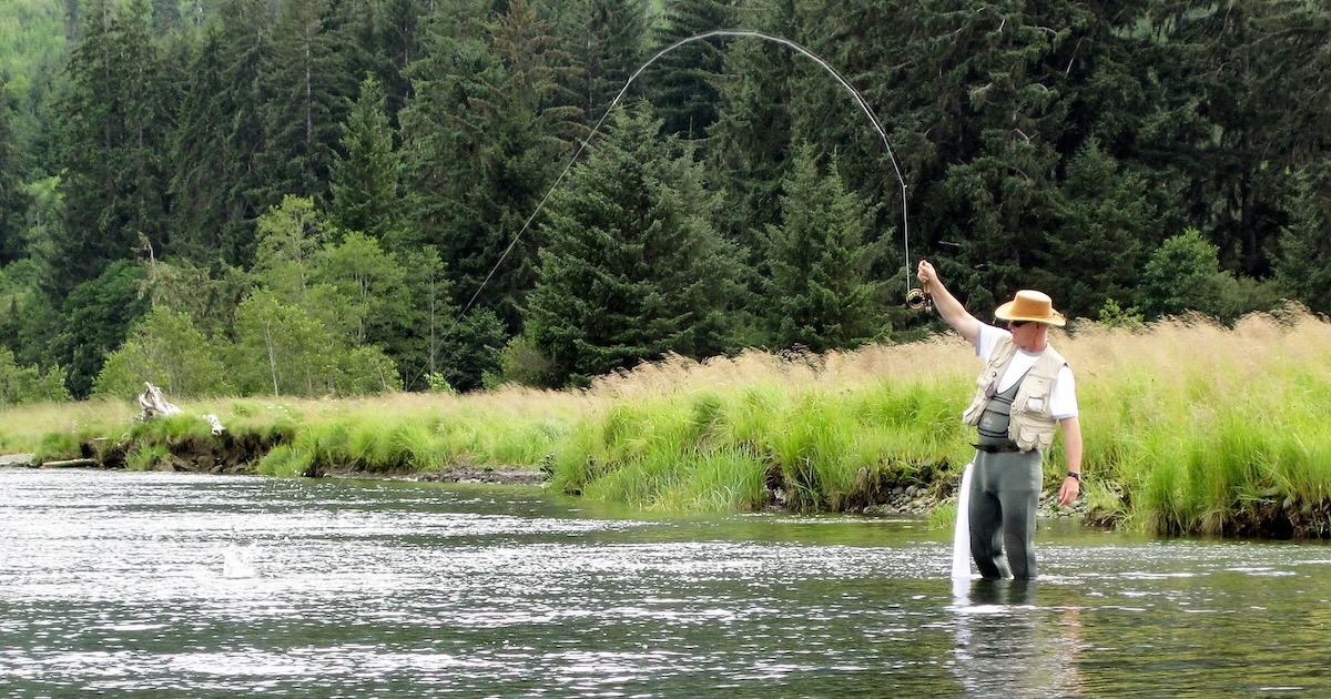 Angler standing in a river, casting a fishing rod with a line extended, surrounded by greenery and trees.