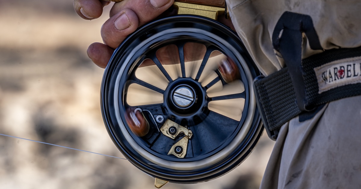 Close-up of a hand holding a centerpin fishing reel with visible spool and handle components.