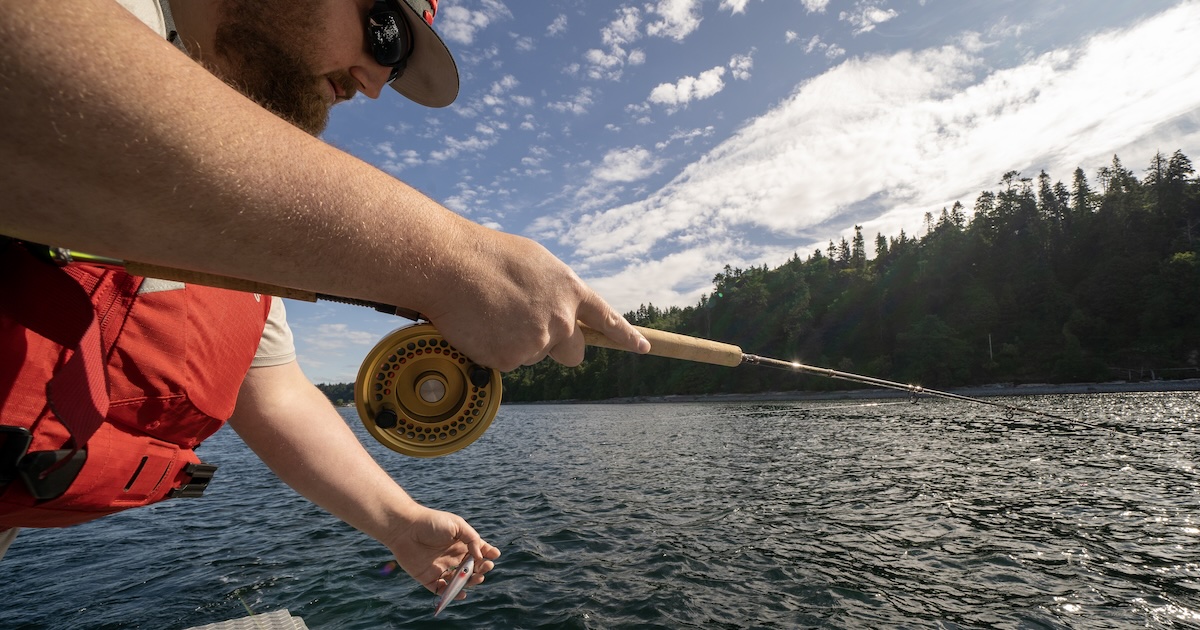 Person in a red life jacket casting a fishing line into the water with a fly rod, surrounded by trees and a cloudy sky.
