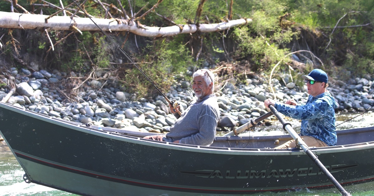 Two individuals in a fishing boat, one holding a fishing rod while the other rows, surrounded by natural scenery.