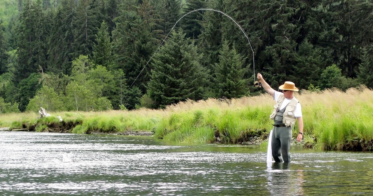 Angler standing in a river, casting a fishing rod with a line extended, surrounded by greenery and trees.