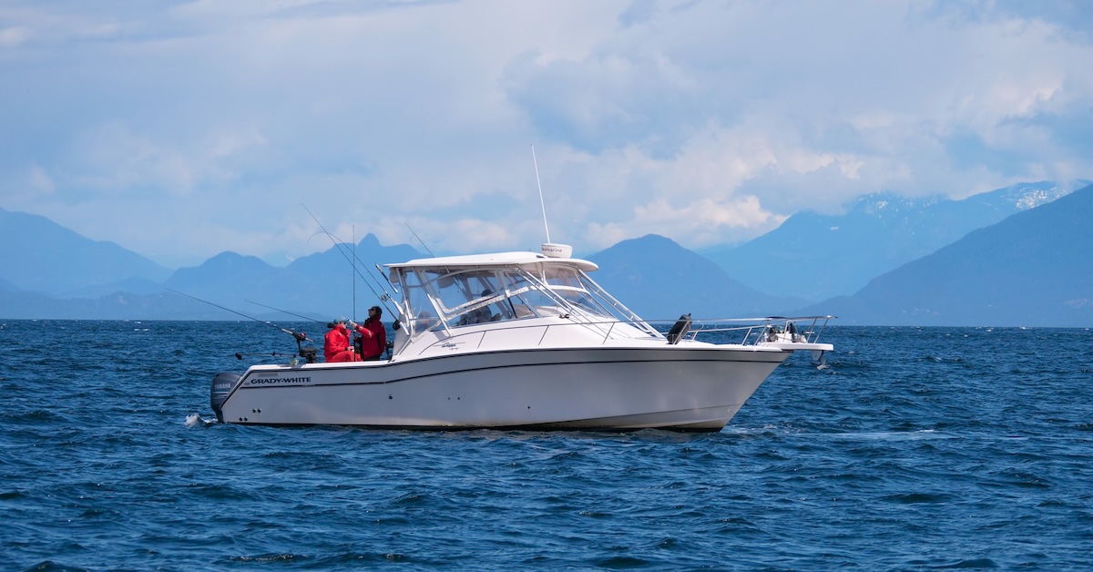 A white Grady-White walkaround fishing boat navigates the deep blue waters near Nanaimo, British Columbia. Two anglers in red waterproof gear are positioned at the stern, tending to multiple trolling lines. The backdrop features the dramatic, layered blue silhouettes of the Coast Mountains under a bright, cloud-dappled sky. Will the Salmon Allocation Policy Change this way of life?