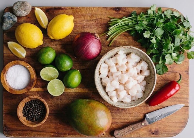 All the fresh ingredients for a dead-simple halibut ceviche, laid out and ready to go: cubed fish, mango, jalapeño, citrus, red onion, and cilantro on a worn wooden cutting board.