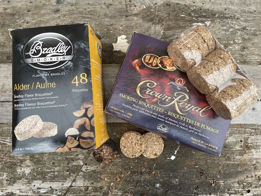 A top-down photograph on a weathered wooden plank surface. On the left is a black-and-yellow cardboard box for Bradley Alder/Aulne smoking bisquettes, specifying '48 Bisquettes' and a weight of '1.6 lb / .726 kg'. To its right is a larger, multi-colored cardboard box for Crown Royal Smoking Bisquettes, featuring text like 'SMOKING BISQUETTES ~ BISQUETTES DE FUMAGE' and 'Parfaites pour utilisation avec toutes marques de fumoir, grill ou barbecue'. Perched on top of the Crown Royal box is a clear shrink-wrapped stack of three original wood bisquettes. On the wooden surface below, there is a burnt, dark charcoal puck, and next to it, two new, unburnt bisquettes. Scattered wood chips are on the surface, and some text (like 'Paquet de 48' and '100% ORGANIQUE - 0 CALORIES') is visible on the Crown Royal box. A small logo with a 'S' in a crown is at the bottom right. Perfect for West Coast Smoked Salmon.