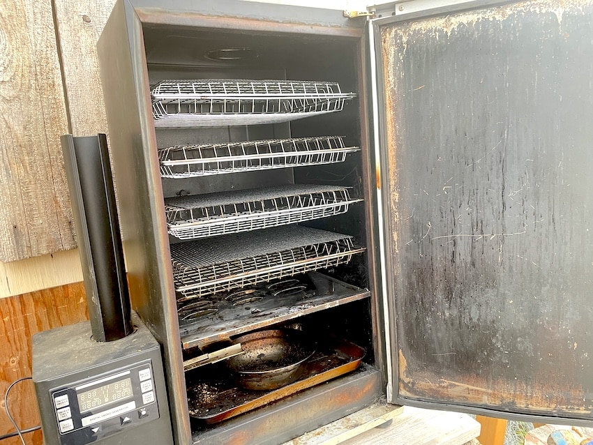 For Smoking Salmon, an open, well-used black Bradley Digital Smoker showing a seasoned interior with four wire smoking racks. The digital smoke generator and bisquette feeder tube are visible on the left. The inside of the door and the drip tray show heavy dark seasoning from frequent use, set against a rustic wooden background.