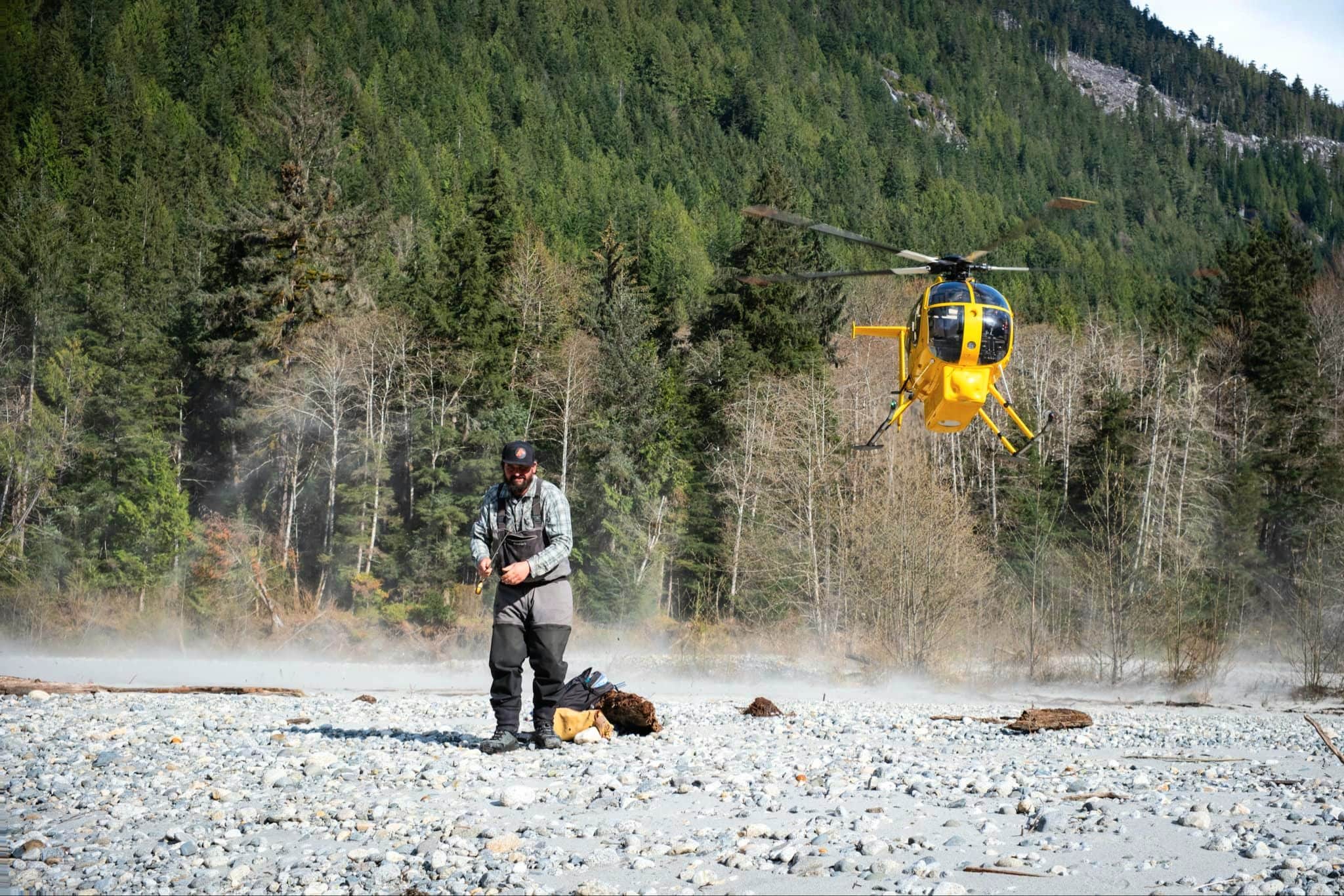 A Stillmaniac Review & Pilgrimage: Fly Fishing for Coastal Cutthroat Trout in Powell River Pat Demeester standing on a misty river gravel bar with a fly rod as a yellow helicopter hovers in the Powell River backcountry